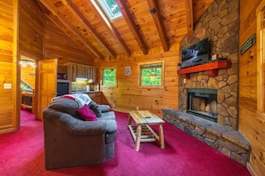 Ein Holzblockhaus-Interieur mit Kamin aus Stein, Holzwänden, einem roten Teppich, einer Couch und einem Fernseher, der über dem Kamin befestigt ist.