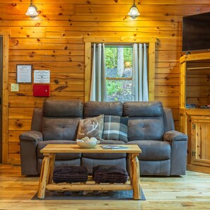 A wooden cabin interior with a sofa, a table with a bowl of snacks, a window with curtains, and a mounted TV.