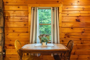 Innenansicht einer Holzhütte mit Esstisch, Stühlen, einem Blumentopf und einem Fenster mit Vorhängen.
