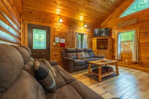 Interior de una cabaña de troncos con un sofá, un sillón reclinable, una mesa de madera y un televisor.
