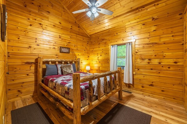 A wooden cabin bedroom with a bed, a ceiling fan, and a window with curtains.