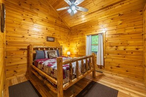 A wooden cabin bedroom with a bed, a ceiling fan, and a window with curtains. - Hunter's Retreat - Cozy Cabin, near Old Man's Cave (Logan)