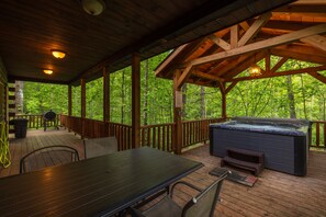A covered outdoor deck with a hot tub, wooden railing, and a table with chairs. - Hunter's Retreat - Cozy Cabin, near Old Man's Cave (Logan)