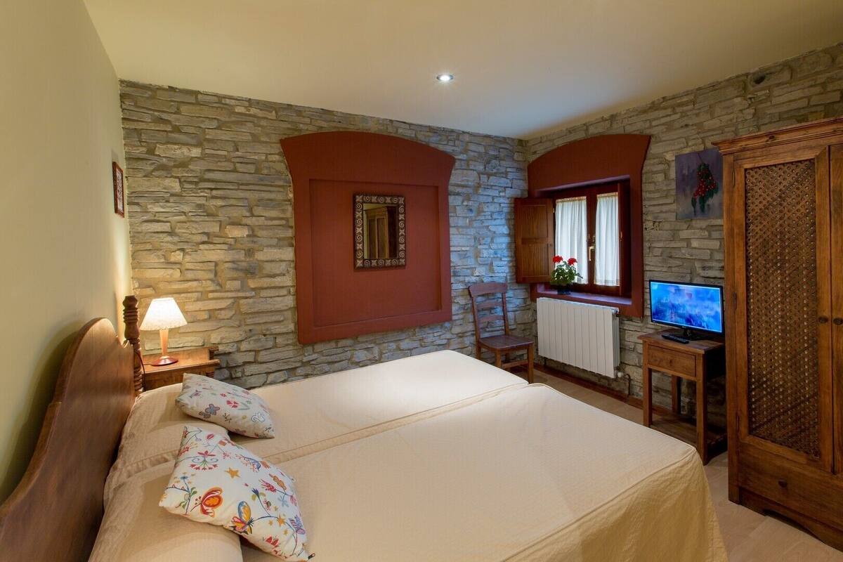 A bedroom with a stone wall, a bed with floral pillows, a wooden nightstand with a lamp, a TV on a wooden stand, and a wardrobe with a woven door.