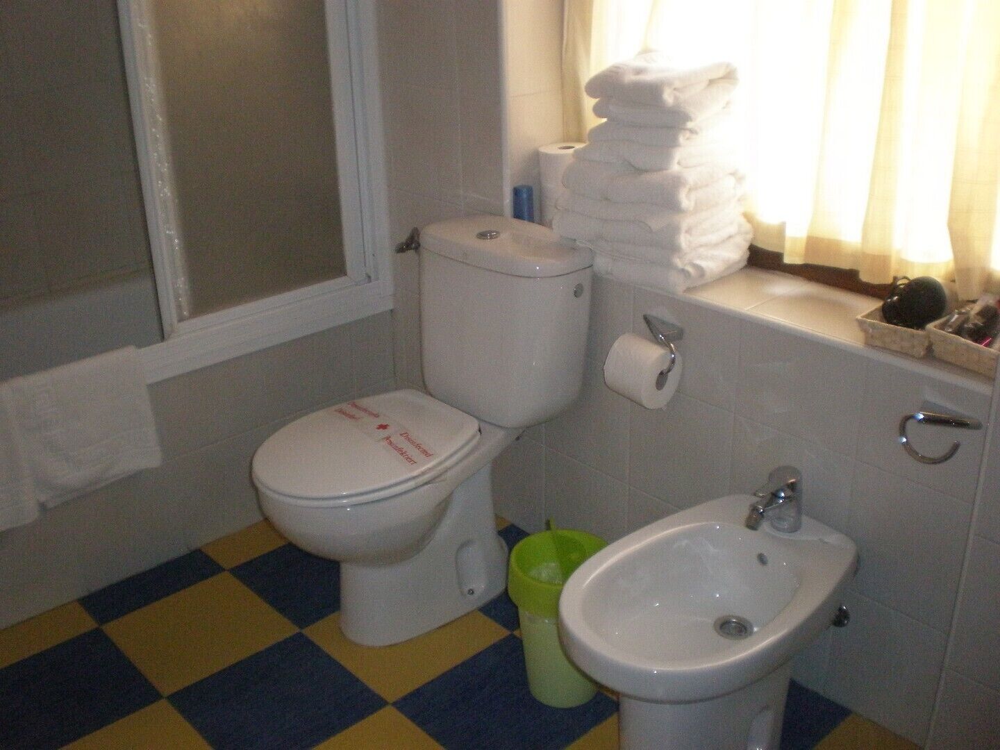 Un baño con inodoro, bidé y lavamanos. Hay una pila de toallas en el mostrador del lavamanos y un cubo de basura verde al lado del inodoro.
