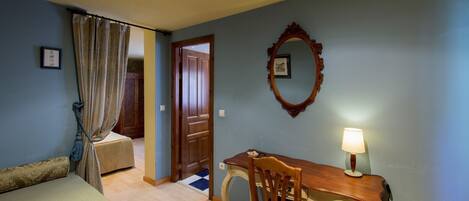 Una habitación con cama, escritorio con silla, lámpara, espejo y puerta que da a otra estancia.