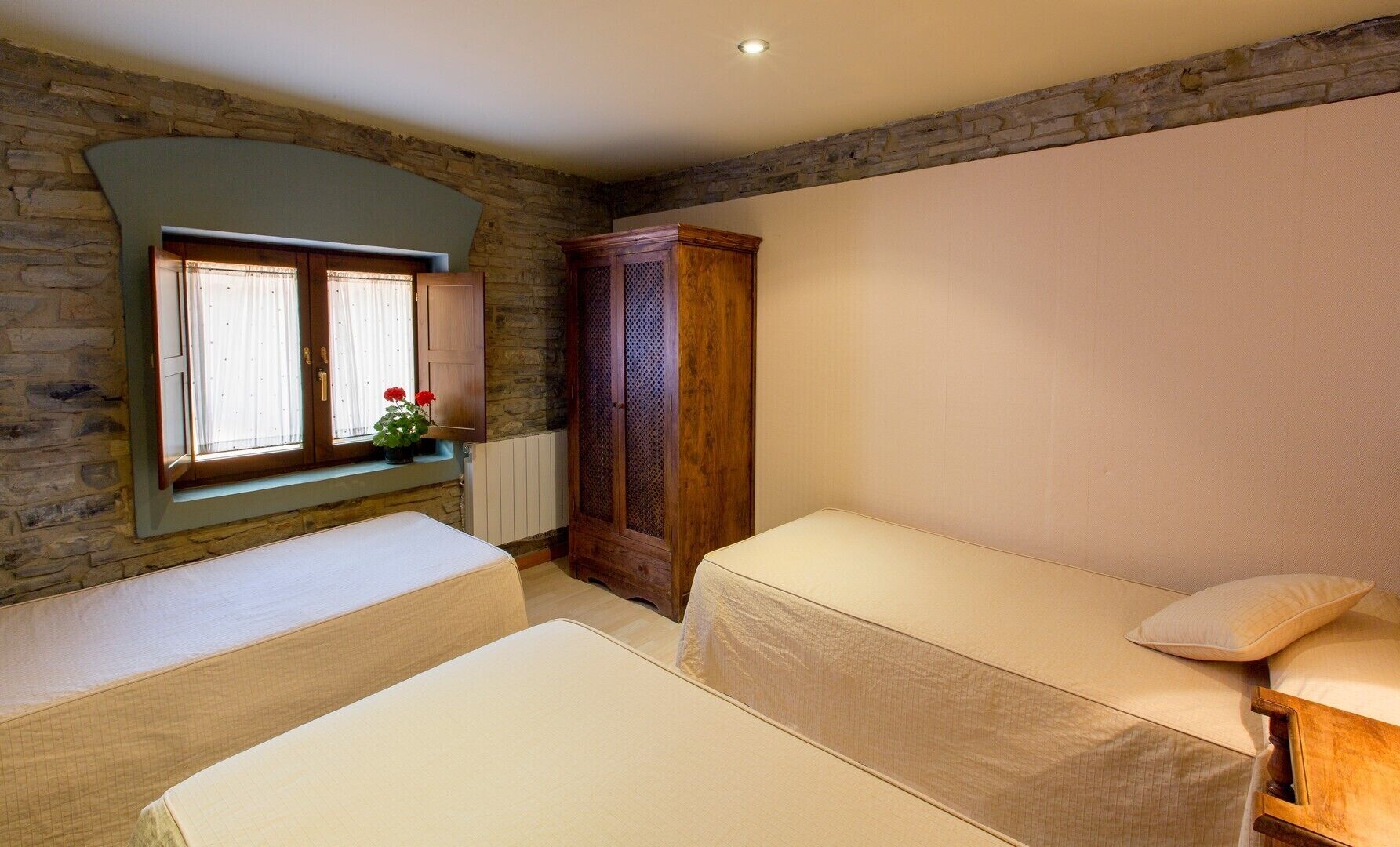 A bedroom with two single beds, a stone wall, a wooden wardrobe, and a window with curtains.