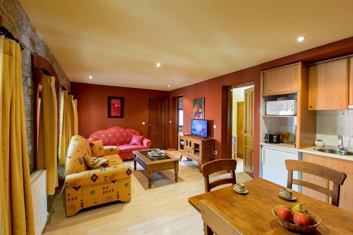 A living room with a red sofa, a wooden coffee table, and a television. There is a kitchen area with wooden cabinets and a microwave.