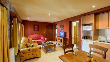 Una sala de estar con un sofá rojo, una mesa de centro de madera y un televisor. Hay una zona de cocina con armarios de madera y un microondas.
