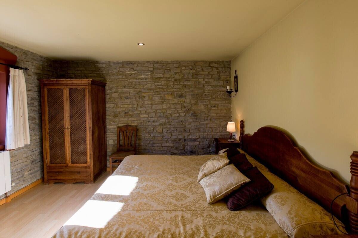 A bedroom with a stone wall, a wooden wardrobe, a wooden bed with patterned bedding, and a nightstand with a lamp.