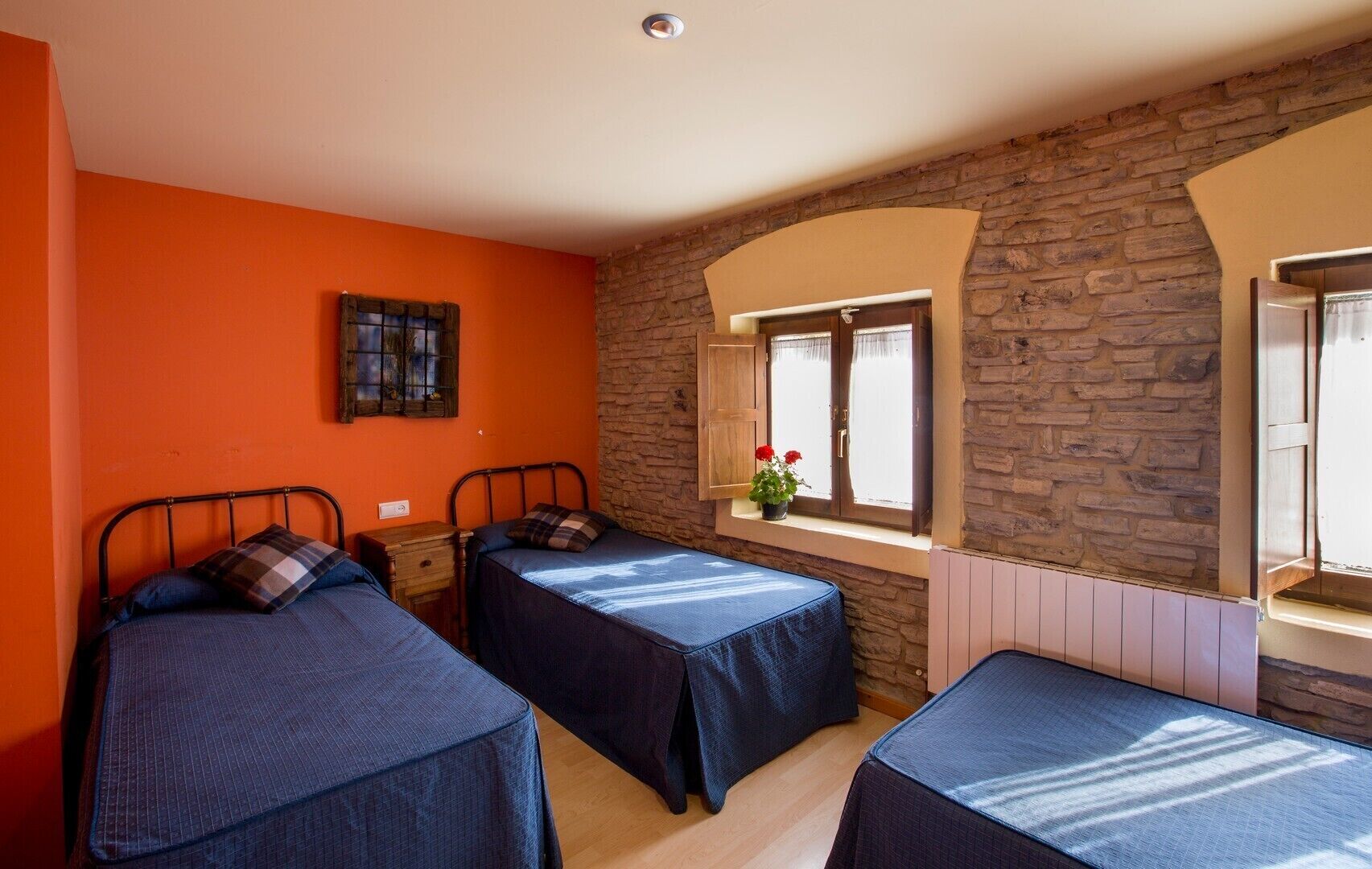 A room with two beds, orange walls, a stone wall, a window with flowers, and a wooden cabinet.