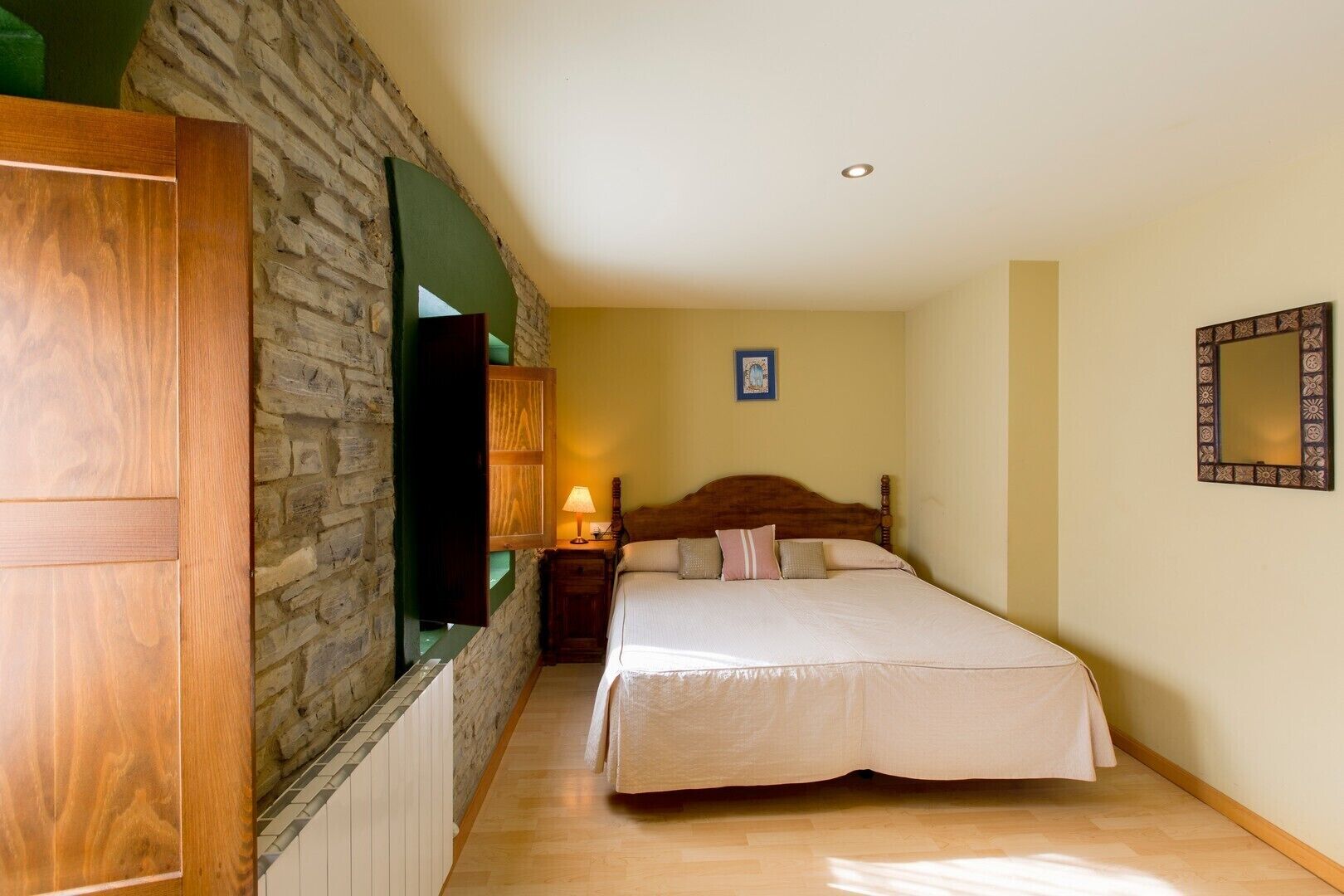 Un dormitorio con pared de piedra, armario de madera, cama con cabecero de madera, mesita de noche con lámpara y espejo en la pared.