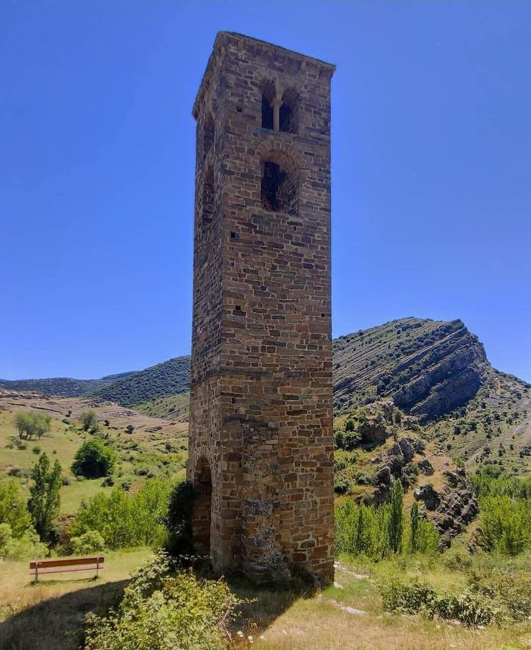 Una torre de piedra alta y esbelta con aberturas arqueadas, con colinas onduladas y un cielo azul despejado de fondo.