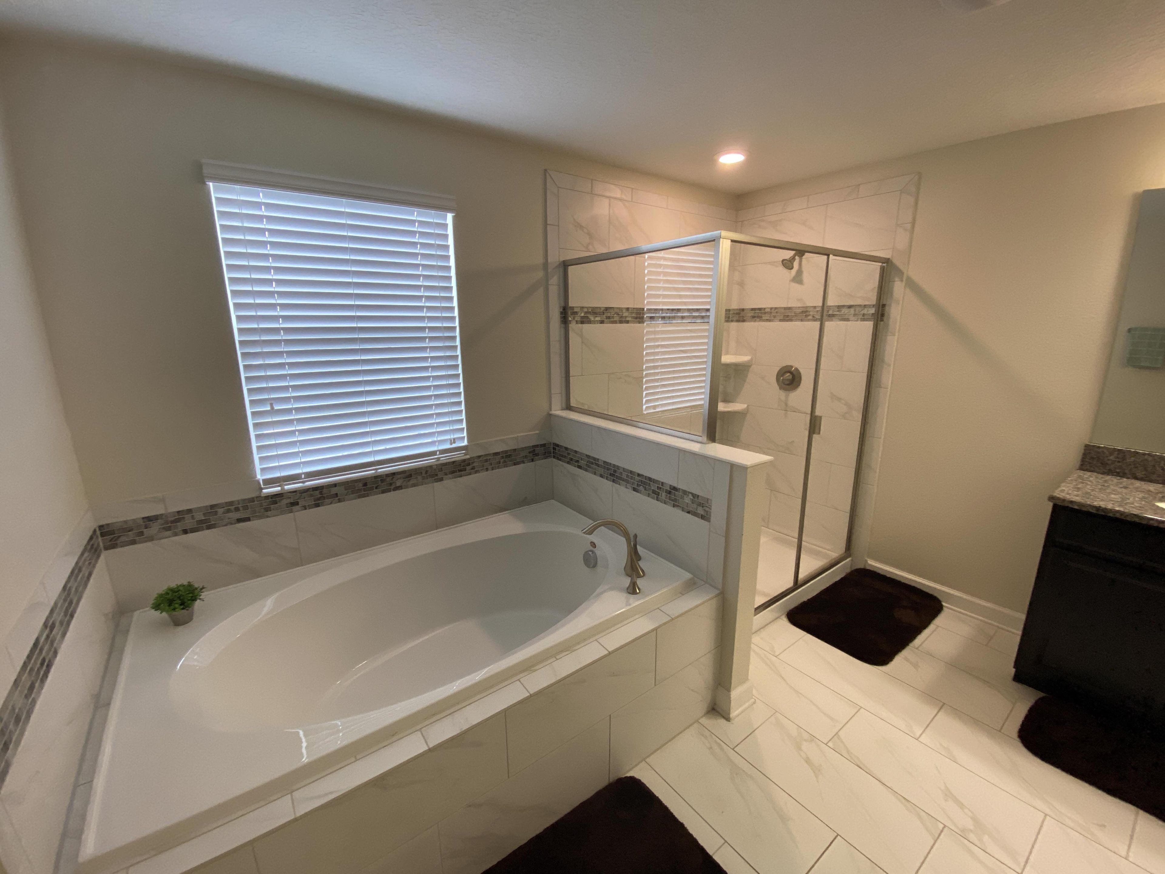 Ein Badezimmer mit einer großen Badewanne, einer gläsernen Dusche und einem Fenster mit Jalousien.