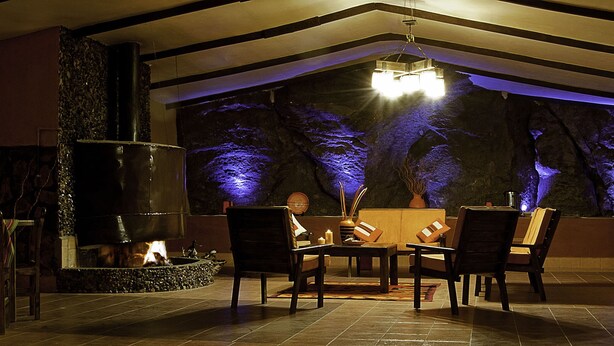 A cozy dining area with a fireplace, stone walls, and a view of trees illuminated by blue lighting.