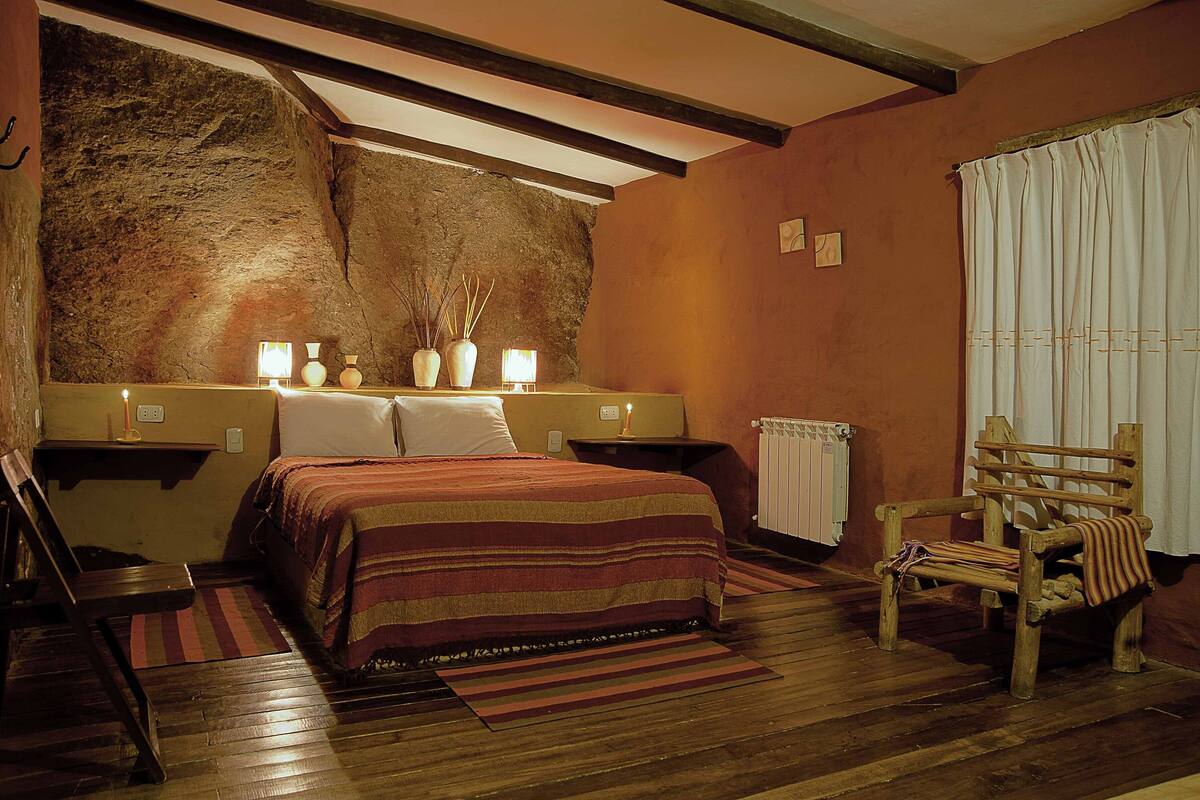 Quarto com cama, mesas de cabeceira, uma cadeira, radiador e parede de pedra.