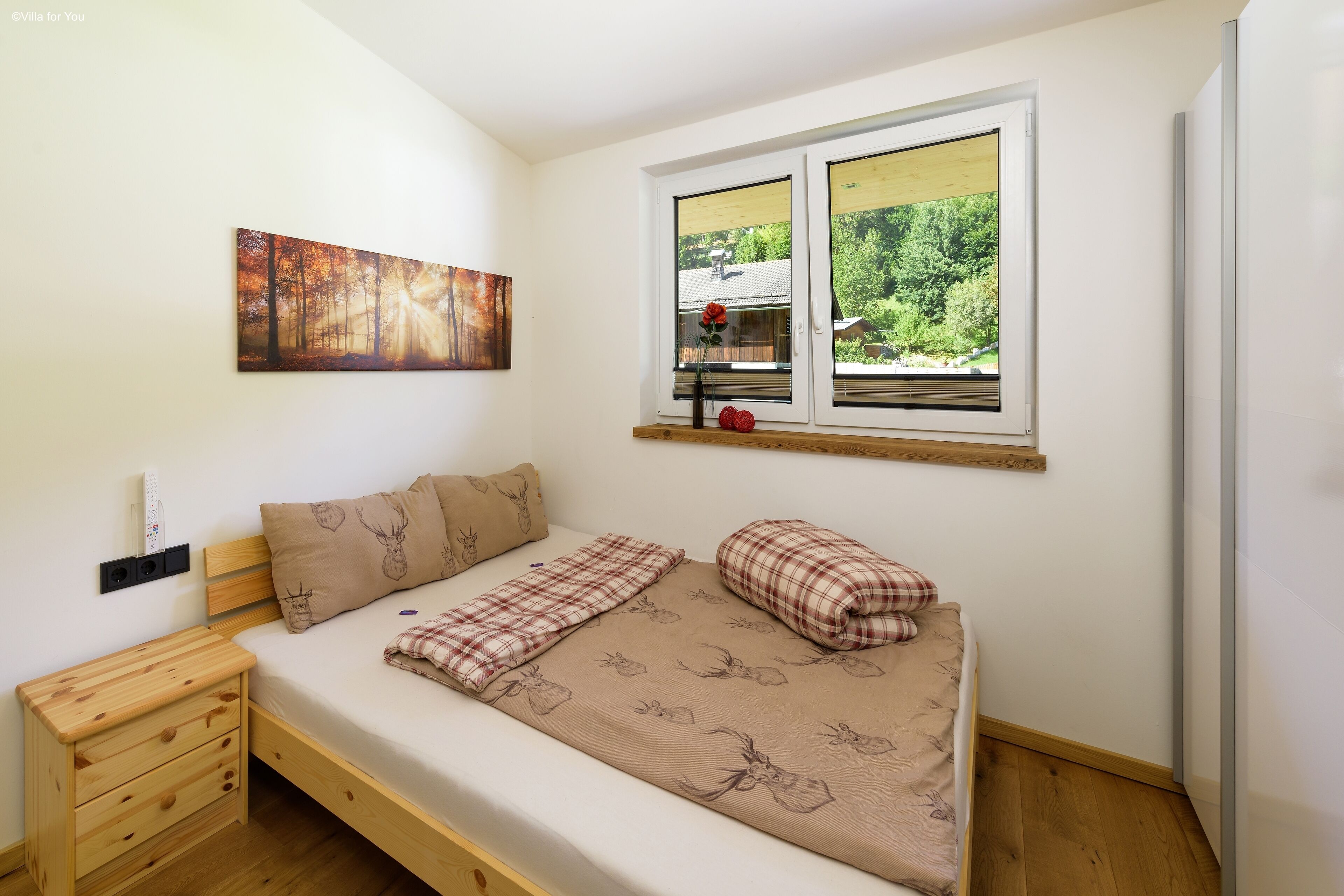 A bedroom with a bed, a nightstand, a window with a view, and a painting on the wall.