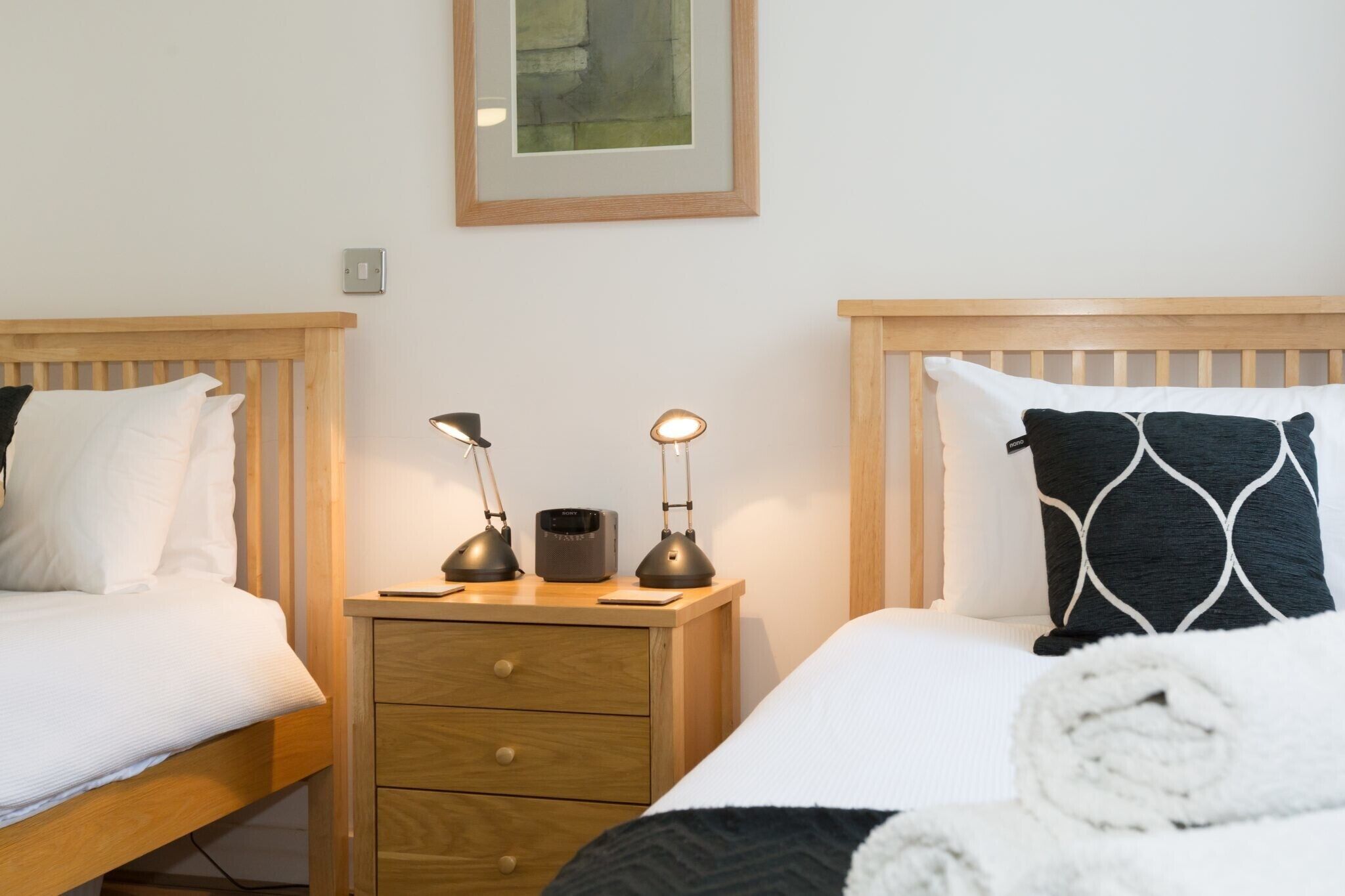 A hotel room with two wooden beds, a nightstand with a lamp, a framed picture on the wall, and a rolled towel on the bed.