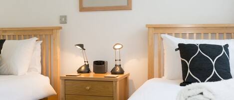 A hotel room with two wooden beds, a nightstand with a lamp, a framed picture on the wall, and a rolled towel on the bed.