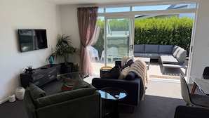A modern living room with a sofa, coffee table, and TV. There is a sliding glass door leading to an outdoor seating area with a couch and a table. - Treasure on Tuscan - Martinborough Holiday Home (Martinborough)