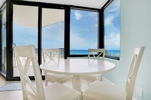 Una mesa redonda blanca con cuatro sillas en una habitación con puertas de vidrio grandes que dan a un balcón con vista al océano.