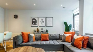 A modern living room with a grey sectional sofa, orange and grey cushions, a wooden coffee table, and a large window providing natural light. - Stunning Modern Apartment in the Heart of Holborn (London)