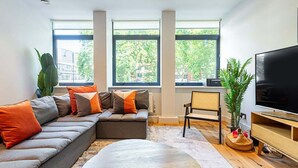 A modern living room with a sectional sofa, a flat-screen TV, and large windows offering a view of trees and buildings. - Stunning Modern Apartment in the Heart of Holborn (London)