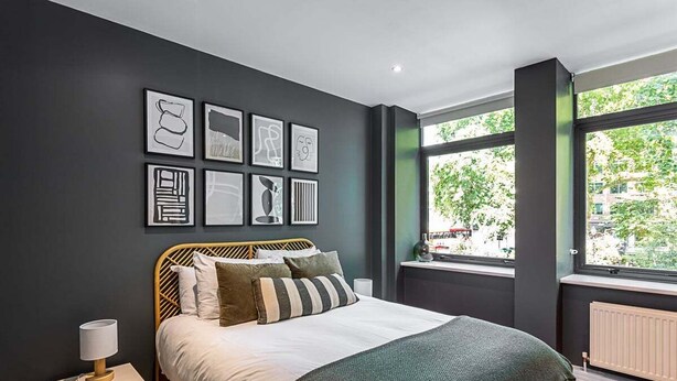 A modern bedroom with a large bed, a bedside table with a lamp, and a window with a view of trees and buildings. - Stunning Modern Apartment in the Heart of Holborn (London)