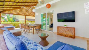 A spacious living area with a large TV, wicker furniture, and a ceiling with wooden beams and hanging lights. - PF42 Excelente Casa 4 Quartos (Mata de São João)