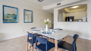 A dining area with a white table, blue chairs, and a vase with flowers. There are two framed artworks on the wall and a kitchen area visible through an archway. - ★Cozy Beach Escape★ 0.9 miles from Clams Pass Beach ! (Naples)