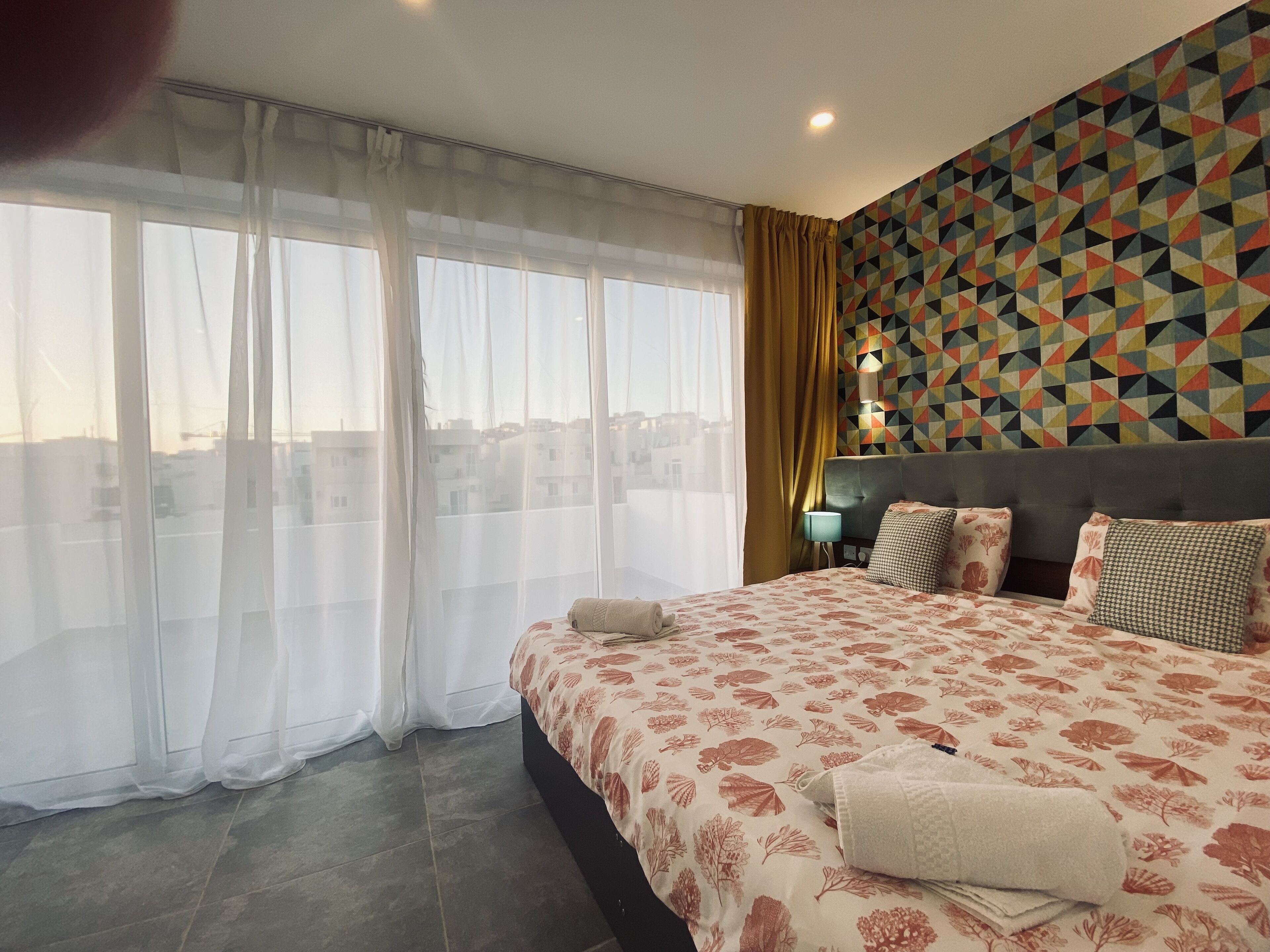 A hotel room with a large bed, a view of the city, and a geometric-patterned wall.