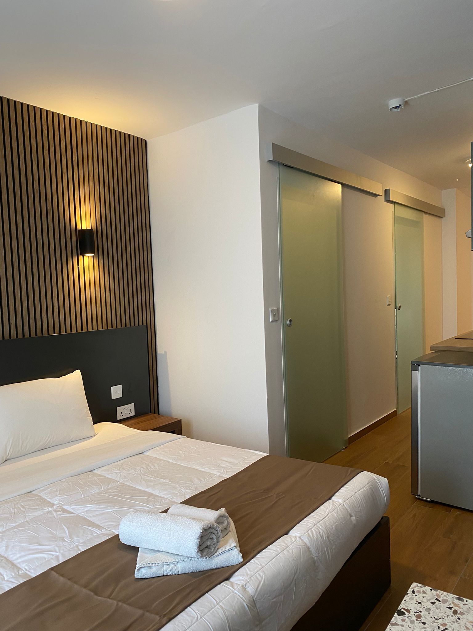 A modern hotel room with a bed, a wooden headboard, a wall-mounted light, a glass door closet, and a kitchenette area.