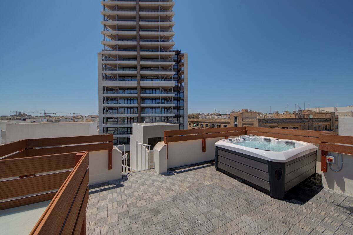 Eine Dachterrasse mit Whirlpool, Holzmöbeln und Blick auf ein hohes Gebäude.