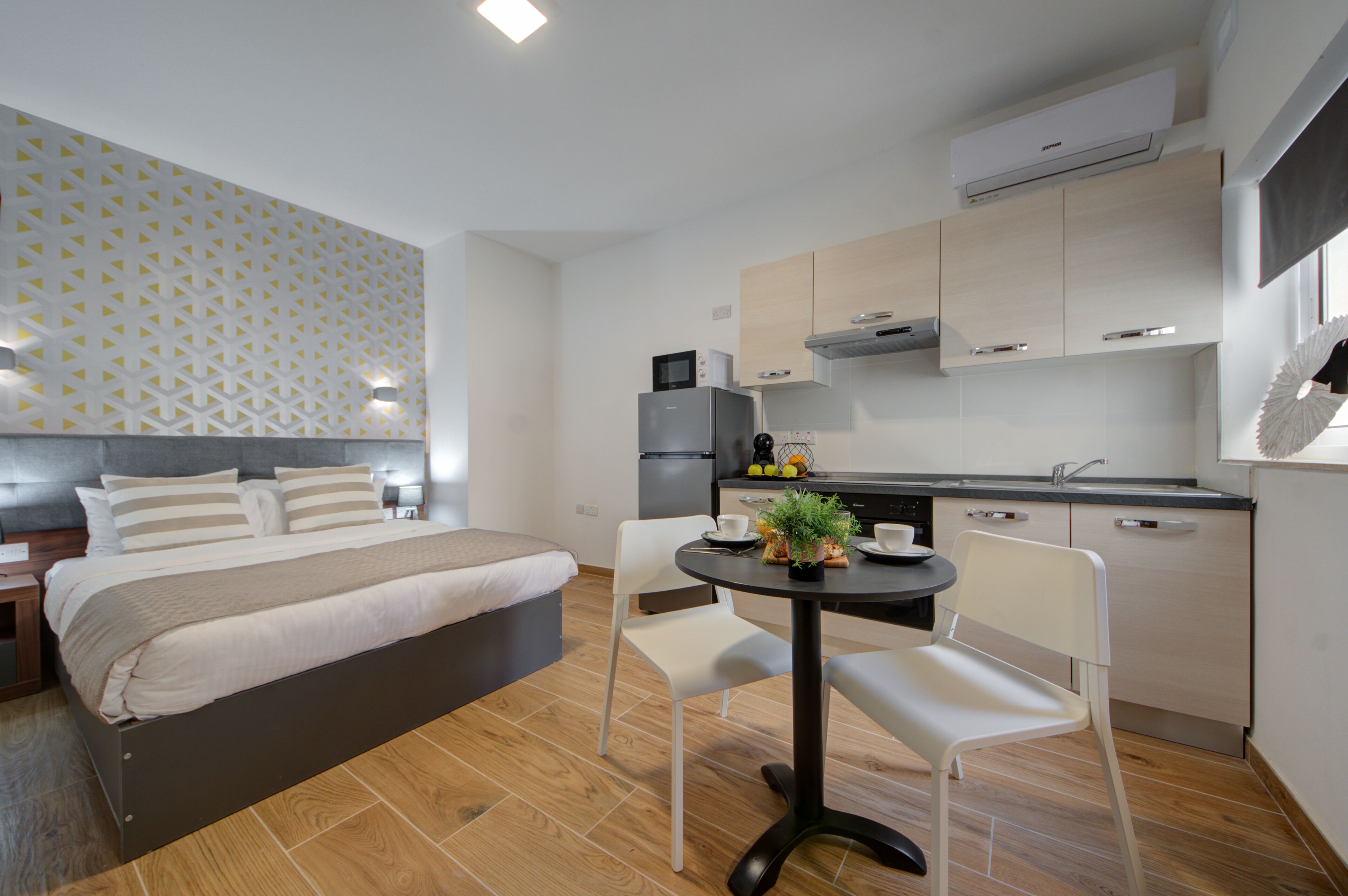 Ein modernes Hotelzimmer mit einem Bett, einem kleinen Esstisch mit Stühlen, einer Küchenzeile und einer Wand mit Muster.