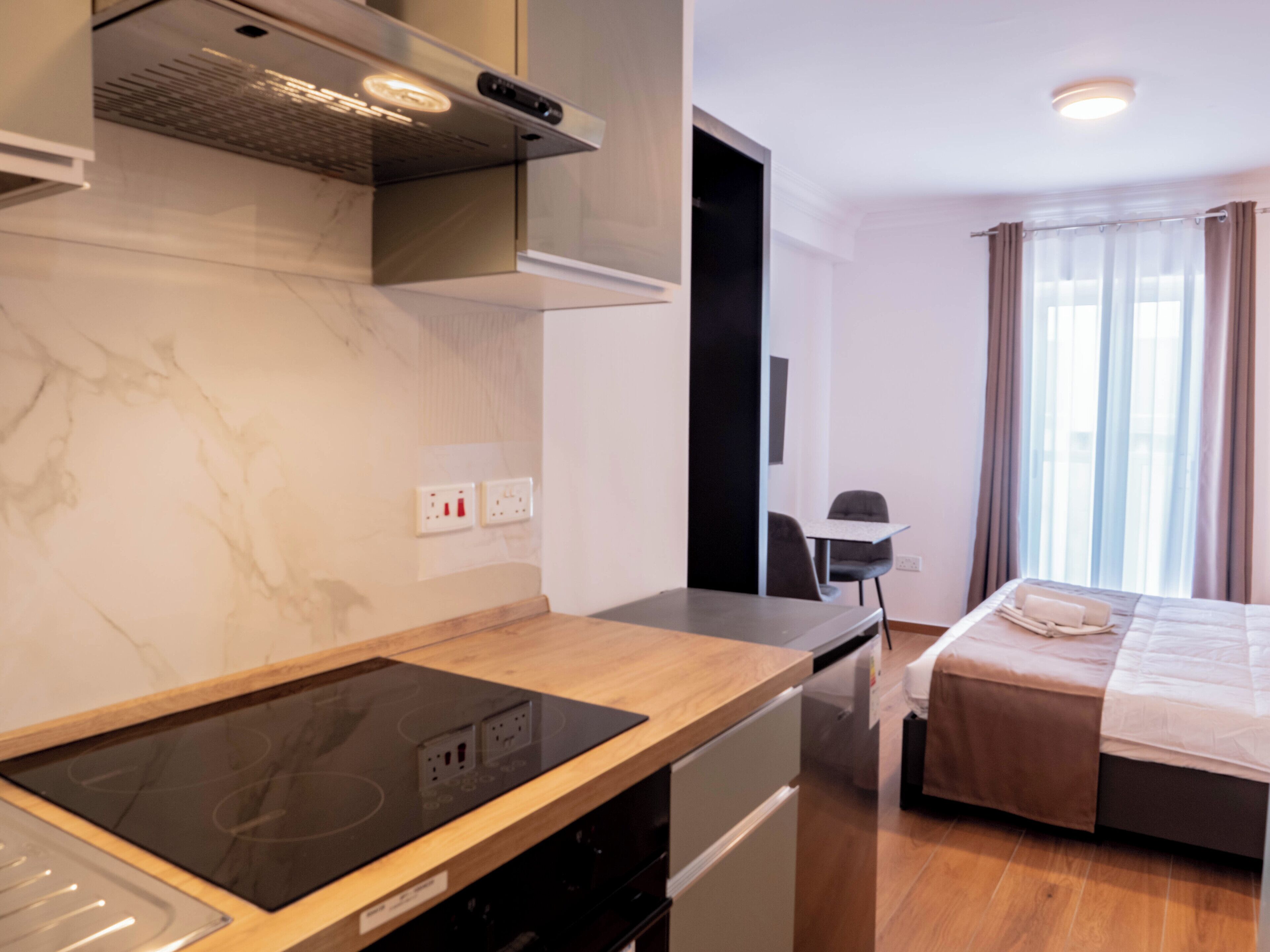 A modern hotel room with a kitchen area featuring a stove, a dining table, and a bed with a view of the city.