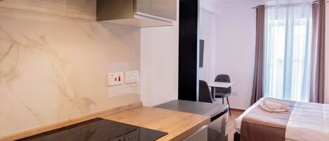 A modern hotel room with a kitchen area featuring a stove, a dining table, and a bed with a view of the city.