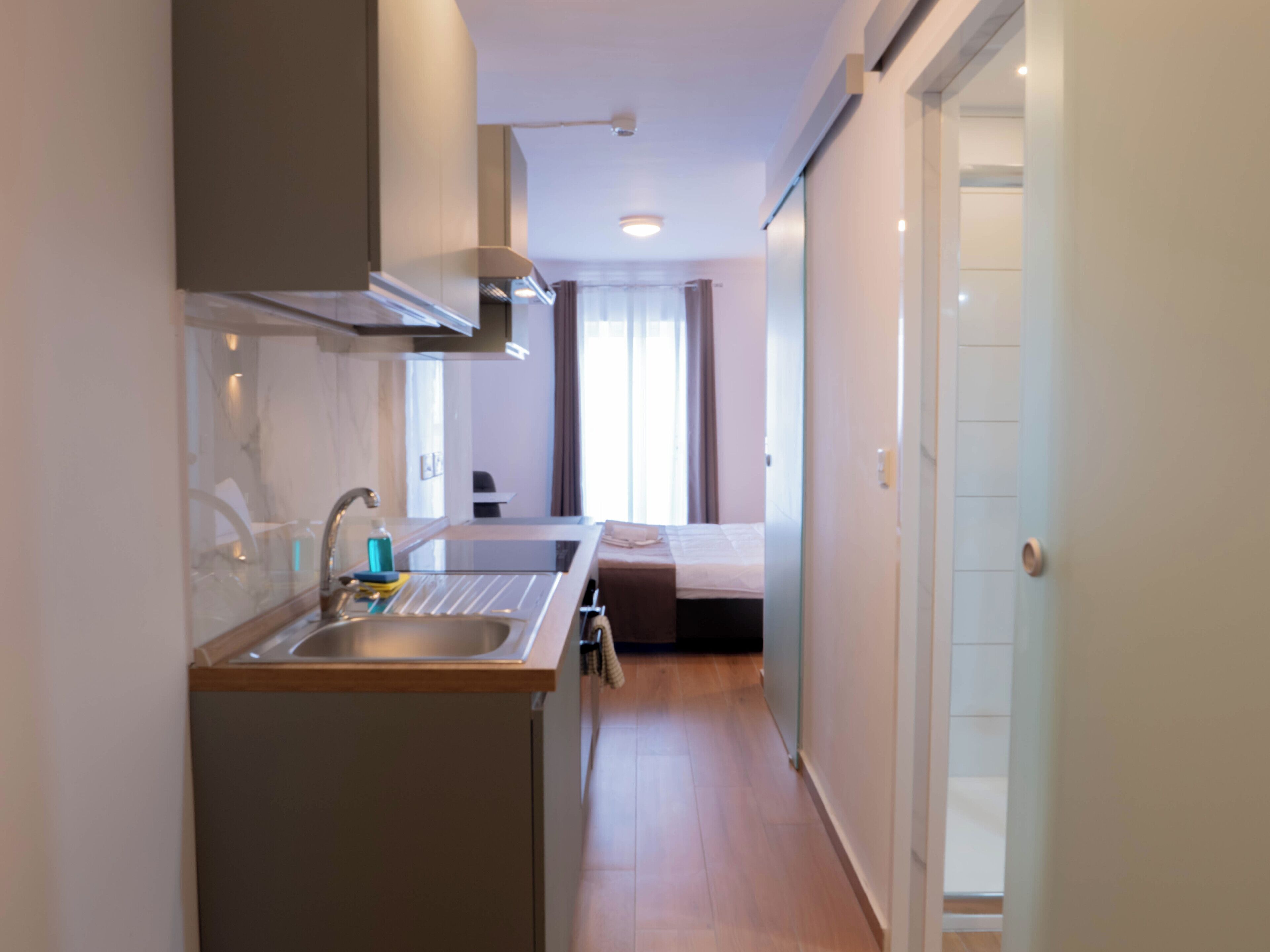 A compact kitchen with a sink, a stove, and a small countertop. A bed is visible in the background, and the room has a sliding door.
