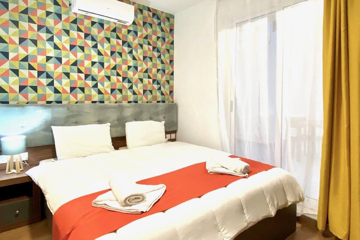 Ein Hotelzimmer mit einem großen Bett, einer Wand mit geometrischem Muster, einem Fenster mit Vorhängen und einem Nachttisch mit Lampe.
