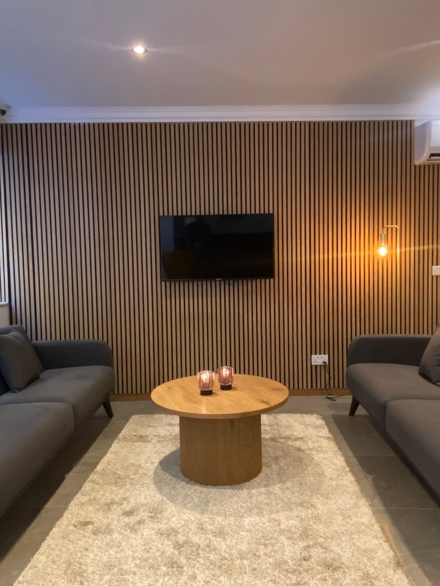 A modern living room with a flat-screen TV mounted on a wooden wall, two sofas, a round coffee table, and a carpeted floor.