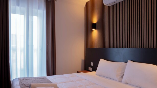 Une chambre d’hôtel avec un grand lit, une tête de lit ornée de lignes verticales, une fenêtre avec des rideaux et un appareil de climatisation fixé au mur.
