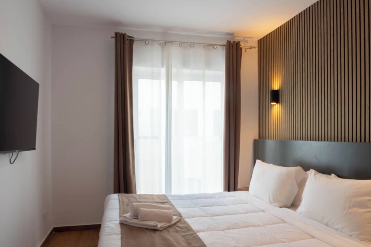 Ein Hotelzimmer mit Bett, Flachbildfernseher, einem Fenster mit Vorhängen und einer Wandleuchte.