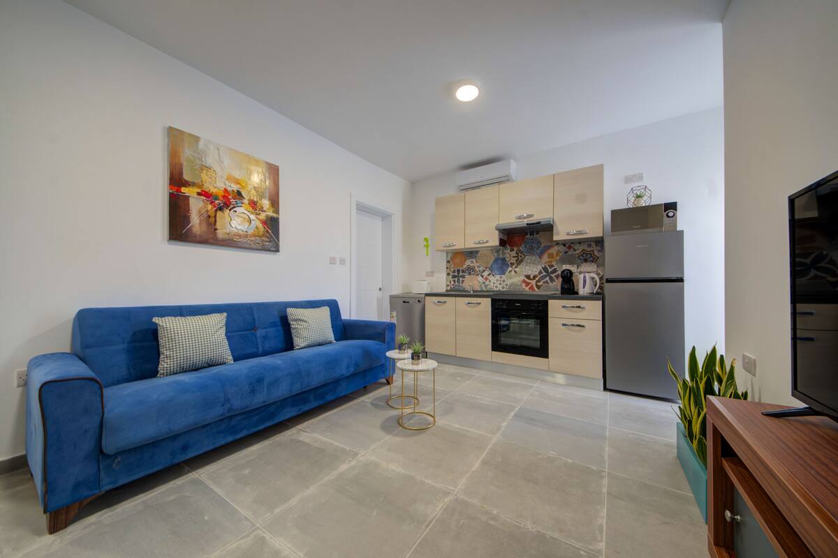 Ein modernes Wohnzimmer mit einem blauen Sofa, einem kleinen runden Tisch und einer Küchenzeile mit Kühlschrank, Herd und Schränken.