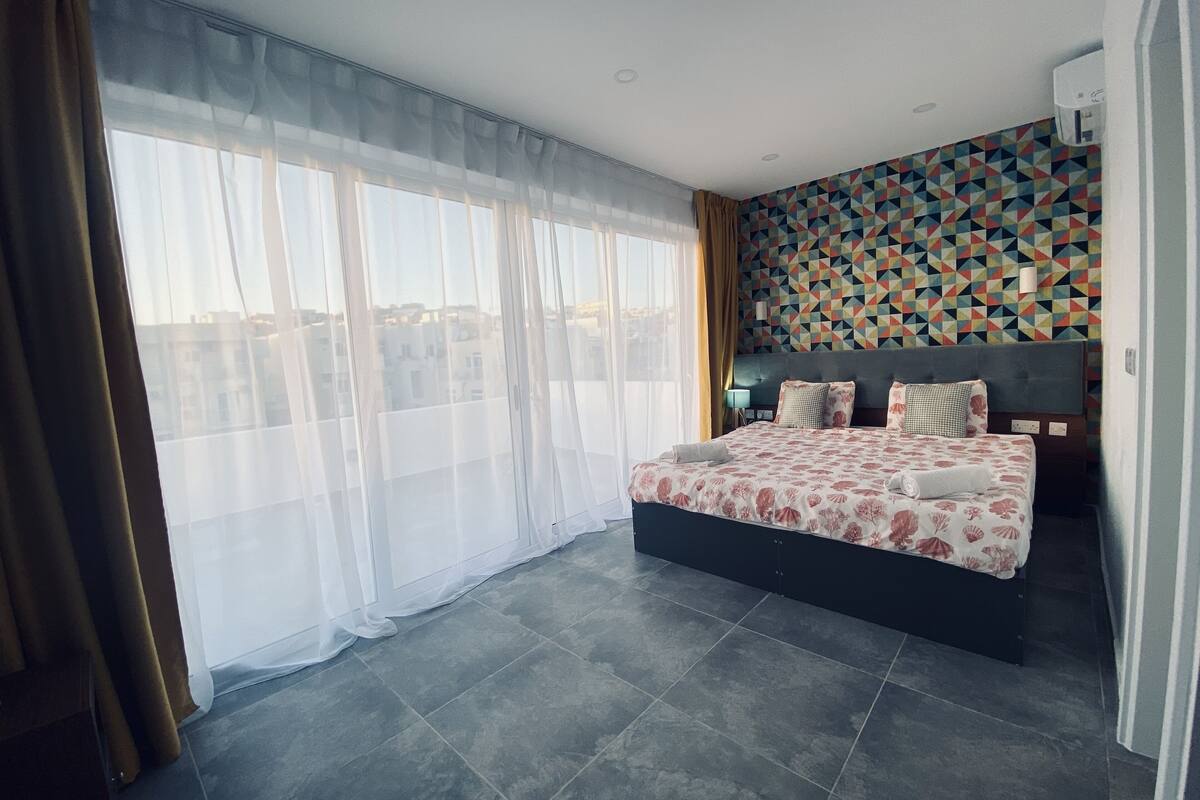 Ein modernes Schlafzimmer mit einem großen Bett, durchscheinenden Vorhängen, einer Wand mit geometrischem Muster und Blick auf die Stadt.