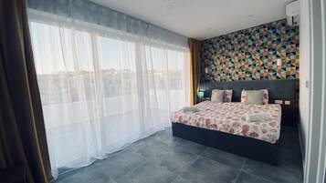 A modern bedroom with a large bed, sheer curtains, a geometric-patterned wall, and a city view.