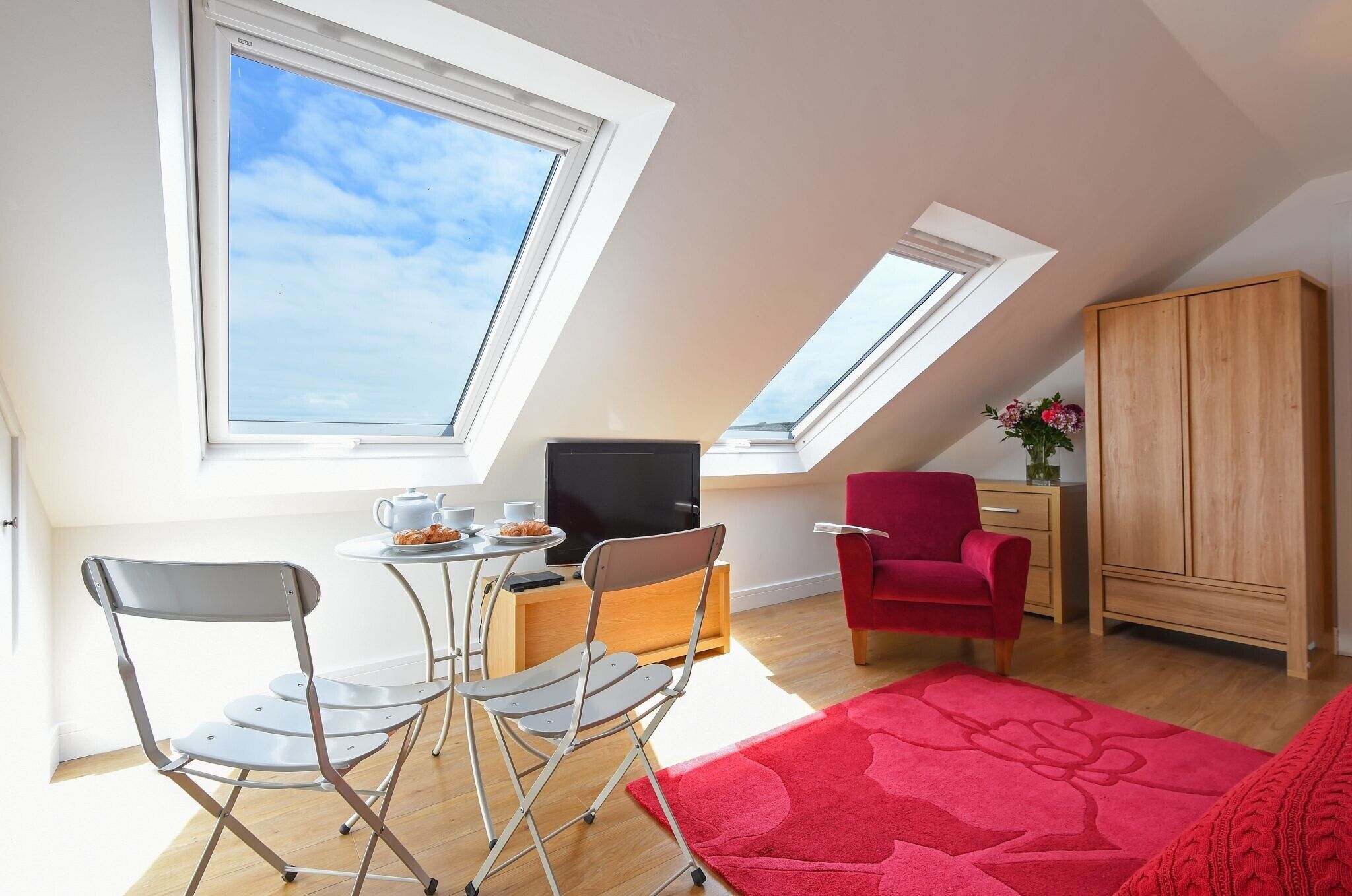 A modern loft apartment with skylights, a dining area with a table and chairs, a red armchair, a wooden cabinet, and a television on a wooden stand.