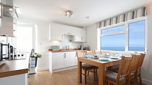 Une cuisine moderne avec des meubles blancs, une table à manger en bois et une vue sur la mer grâce à de grandes fenêtres.