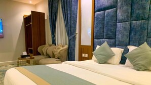 Quarto de hotel com uma cama grande, um guarda-roupa, um espaço de estar com uma cadeira e um banquinho, e uma televisão na parede.