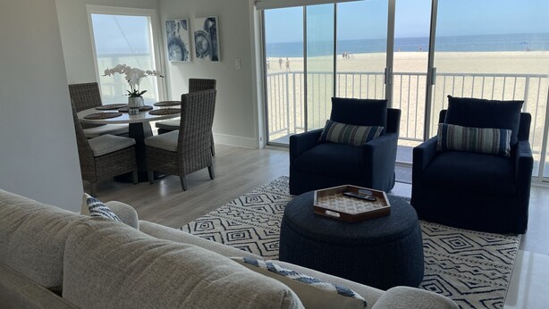 Ein großzügiger Wohnbereich mit einem großen Fenster mit Blick auf einen Strand, ausgestattet mit einer Couch, zwei Sesseln und einem Esstisch mit Stühlen.