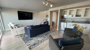 Ein modernes Wohnzimmer mit einem dunkelblauen Sofa, einem weißen Couchtisch und einem Korbstuhl mit Blumen in einer Vase. Eine Treppe führt zu einer Küche mit weißen Schränken und einem Kühlschrank.