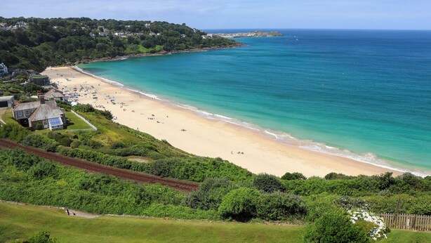 Una playa costera con orilla arenosa, mar azul cristalino y colinas al fondo.